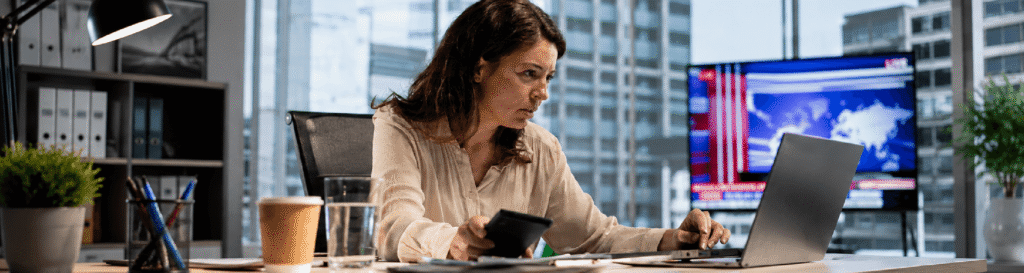 Mulher em um escritório, olhando para a tela e segurando uma caneta e fazendo anotações em um papel