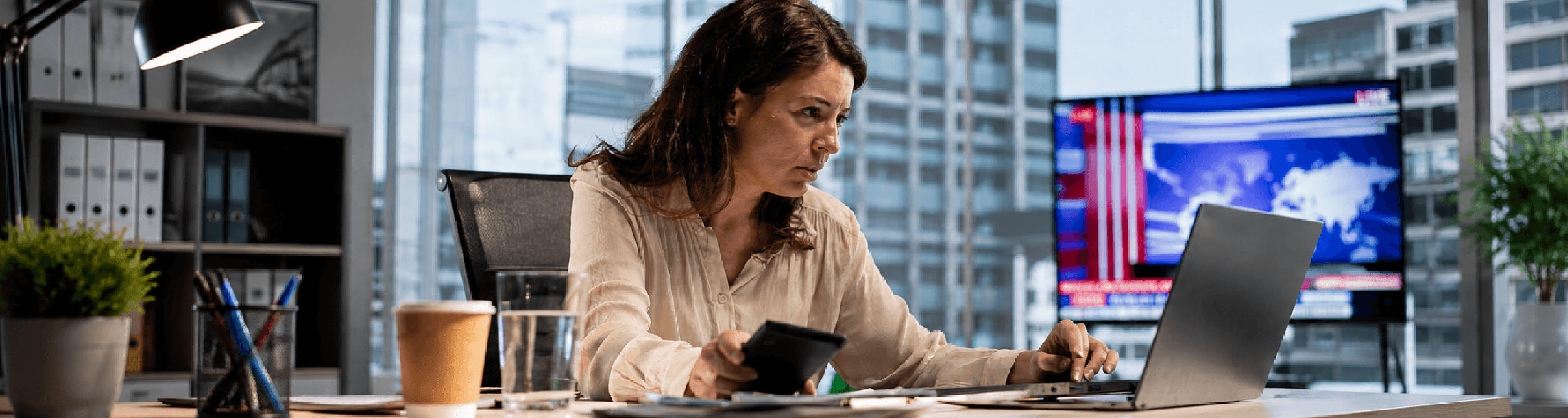 Mulher em um escritório, olhando para a tela e segurando uma caneta e fazendo anotações em um papel